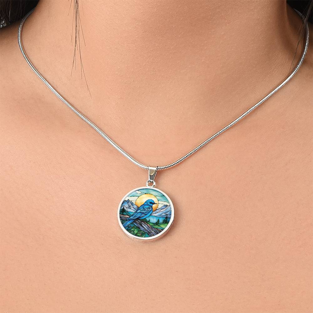 Woman wearing an Elegant silver pendant Blue Bird necklace. Colors blue bird and water, yellow sun, green trees and grass, brown branch, blue and white sky/ mountains. This jewelry offers personalization with an engraving option for a name.