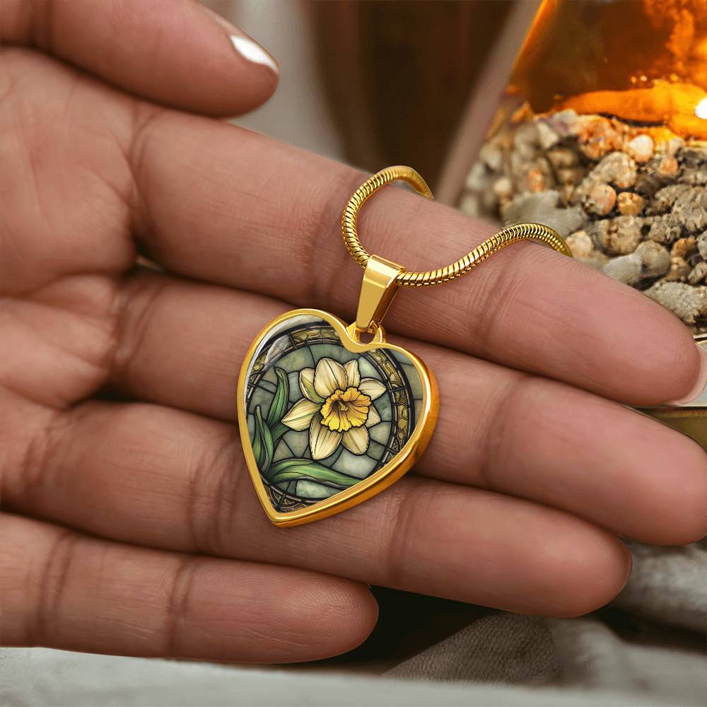 Woman holding an Elegant gold pendant featuring a vivid Daffodil Birth Flower Necklace, March birth month flower, set against a Green & Tan backdrop. This jewelry offers personalization with an engraving option for a name.
