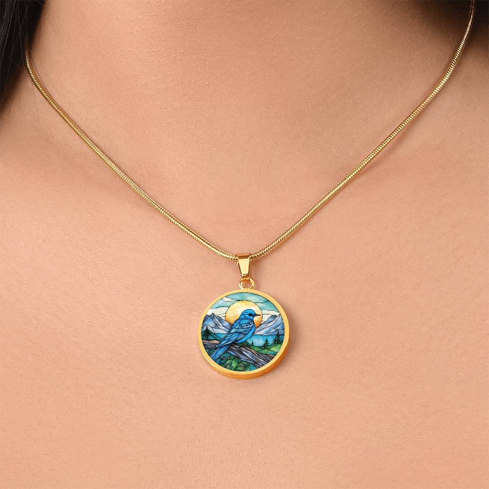 Woman wearing an Elegant gold pendant Blue Bird necklace. Colors blue bird and water, yellow sun, green trees and grass, brown branch, blue and white sky/ mountains. This jewelry offers personalization with an engraving option for a name.