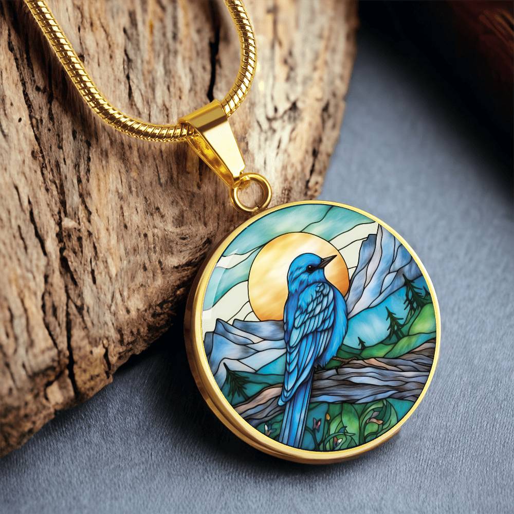 an Elegant gold pendant Blue Bird necklace. Colors blue bird and water, yellow sun, green trees and grass, brown branch, blue and white sky/ mountains. This jewelry offers personalization with an engraving option for a name.