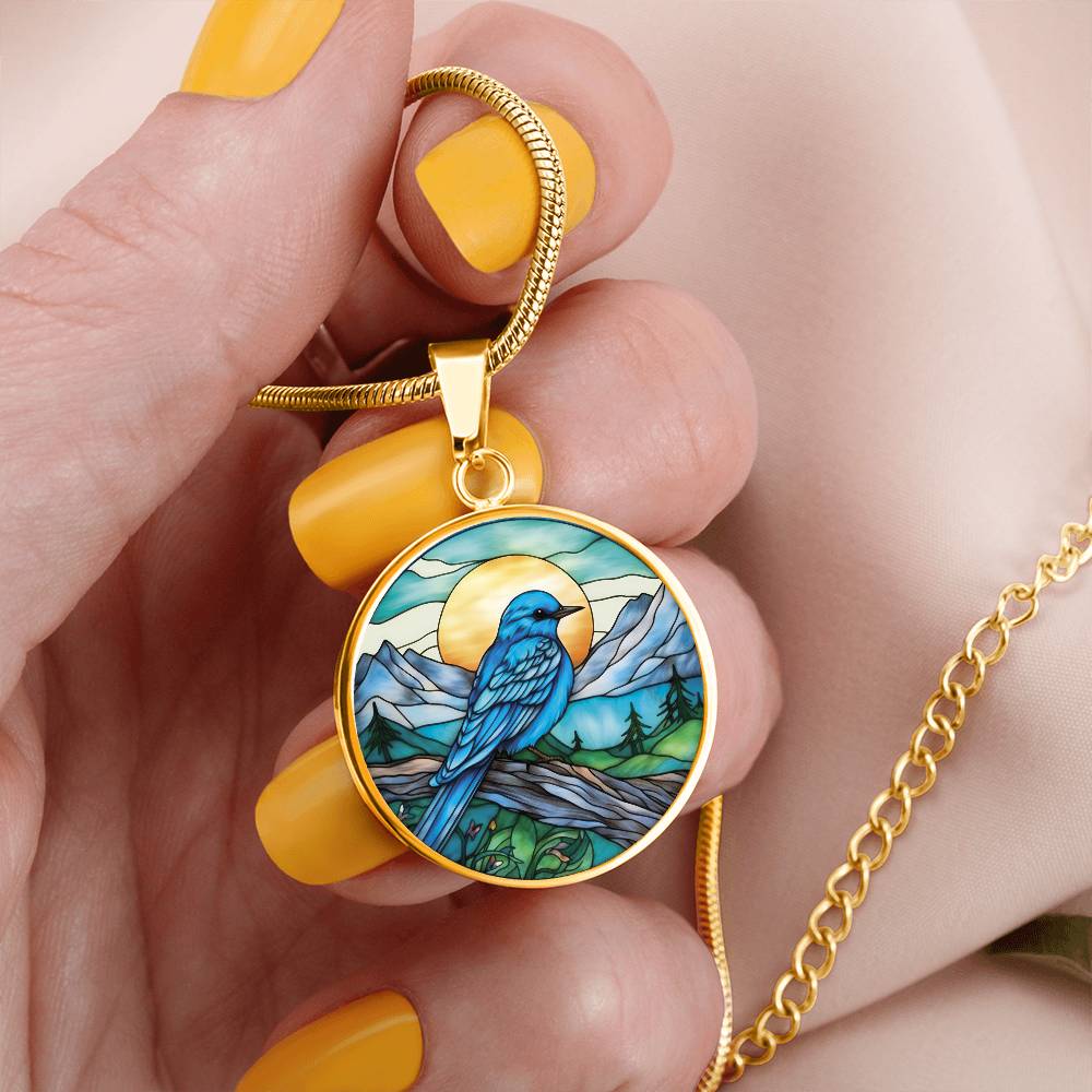 Woman holding an Elegant gold pendant Blue Bird necklace. Colors blue bird and water, yellow sun, green trees and grass, brown branch, blue and white sky/ mountains. This jewelry offers personalization with an engraving option for a name.