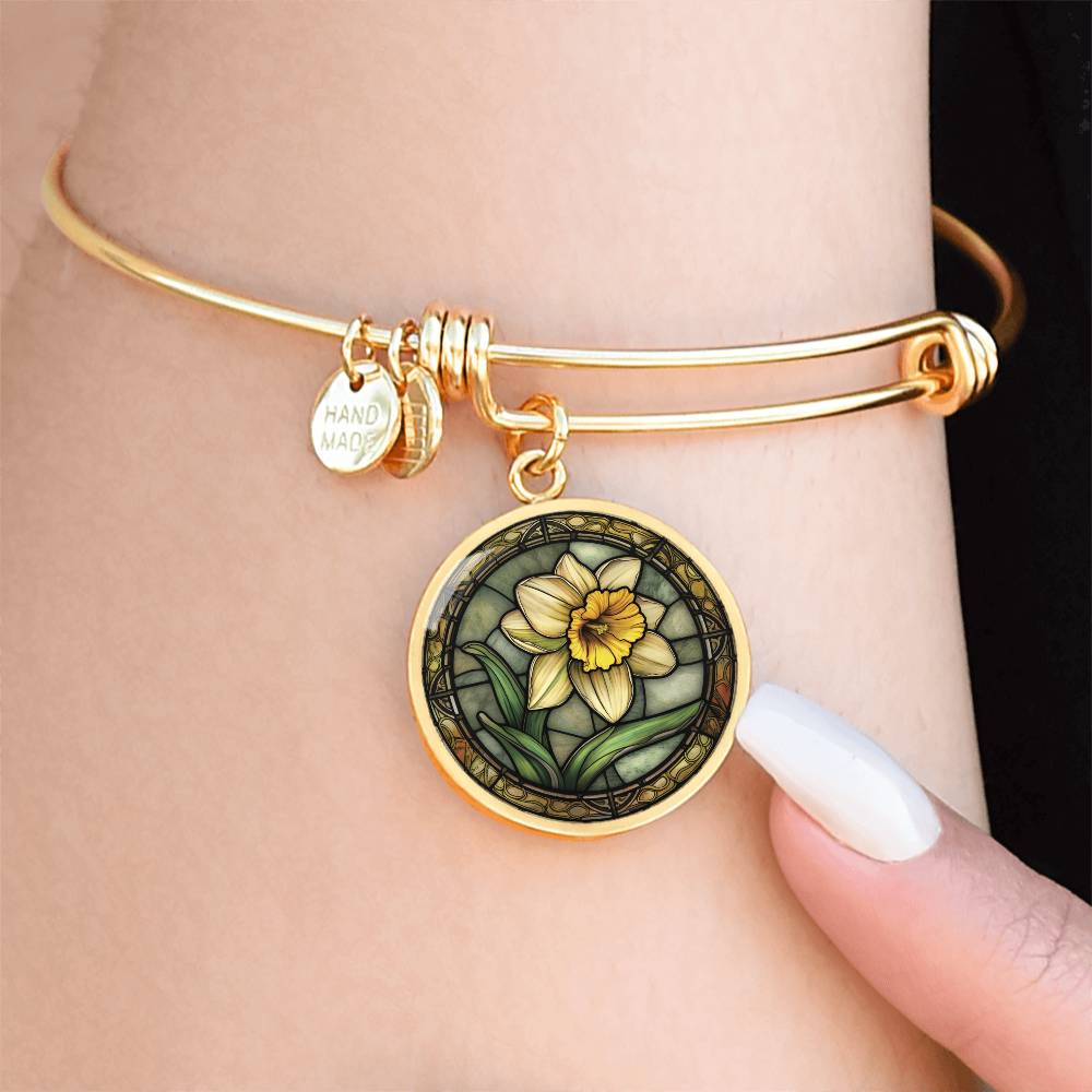 Woman wearing an Elegant gold pendant bracelet featuring a vivid Yellow Daffodil with green grass, set against a tan backdrop with a brown border. This jewelry offers personalization with an engraving option for a name.