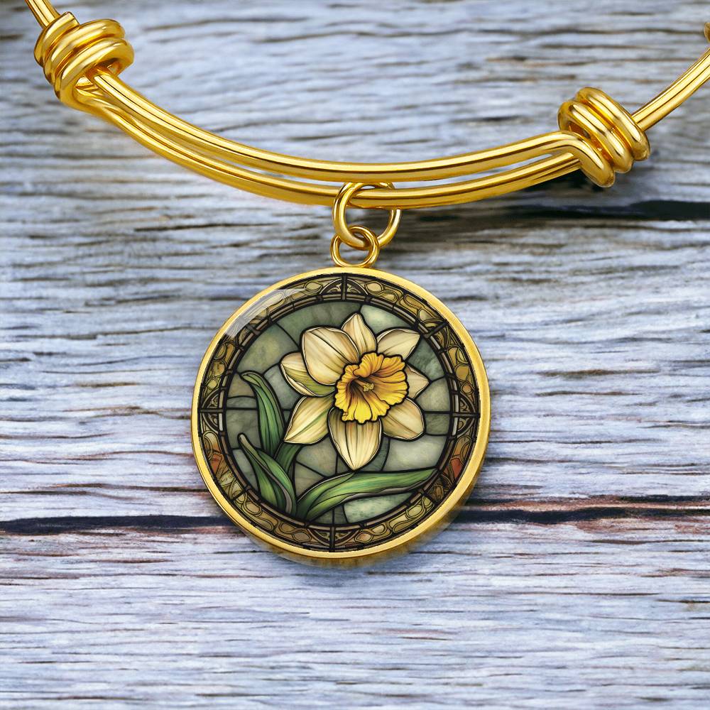an Elegant gold pendant bracelet featuring a vivid Yellow Daffodil with green grass, set against a tan backdrop with a brown border. This jewelry offers personalization with an engraving option for a name.