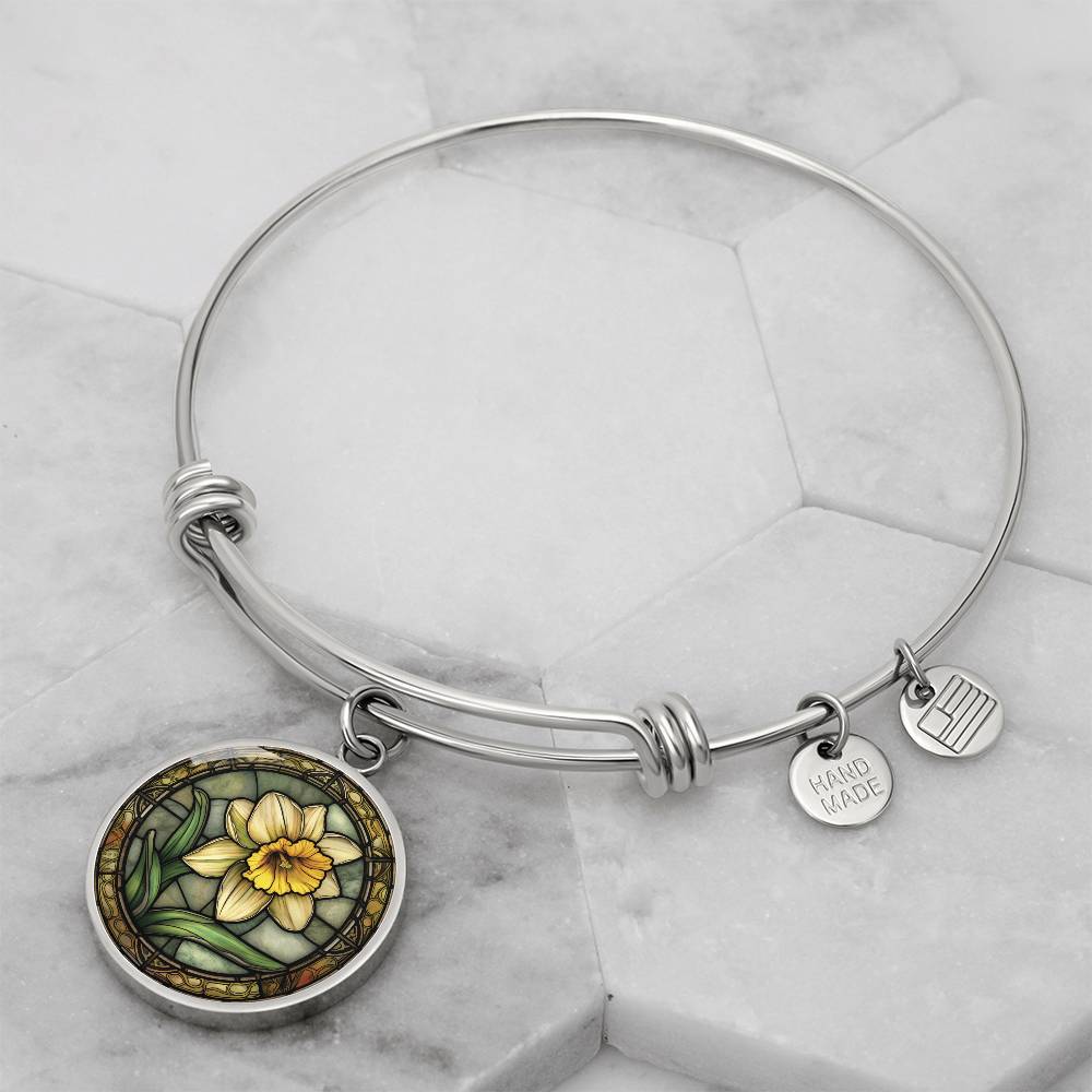an Elegant silver pendant bracelet featuring a vivid Yellow Daffodil with green grass, set against a tan backdrop with a brown border. This jewelry offers personalization with an engraving option for a name.