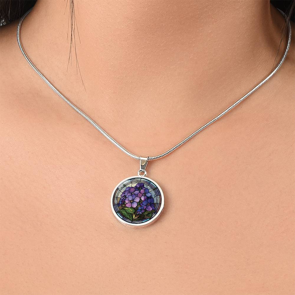 Woman wearing an Elegant silver pendant African Violet Birth Flower Necklace. Violet and purple flowers, green grass, blue, purple, brown background. This jewelry offers personalization with an engraving option for a name.