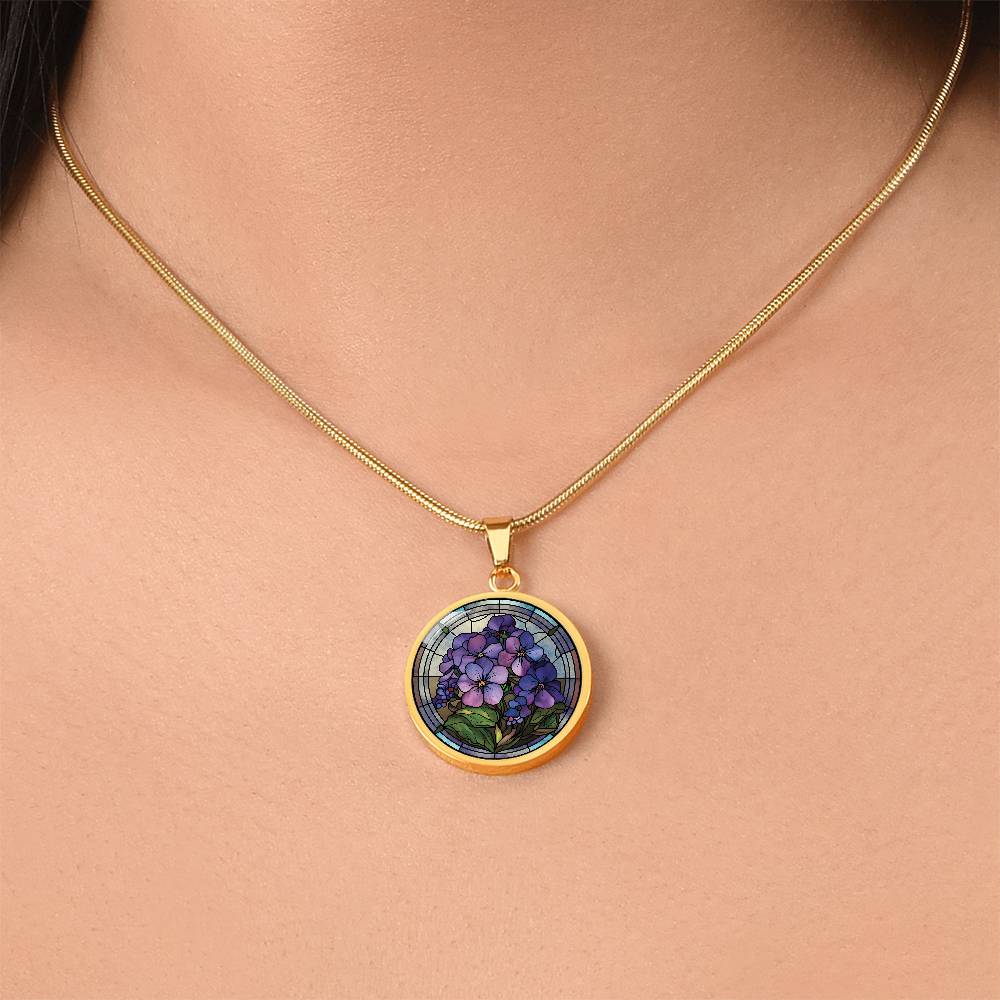 Woman wearing an Elegant gold pendant African Violet Birth Flower Necklace. Violet and purple flowers, green grass, blue, purple, brown background. This jewelry offers personalization with an engraving option for a name.