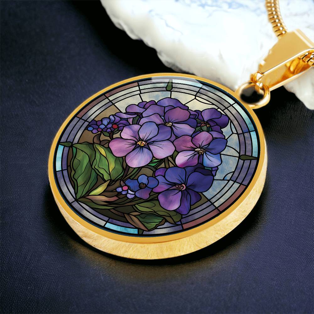 an Elegant gold pendant African Violet Birth Flower Necklace. Violet and purple flowers, green grass, blue, purple, brown background. This jewelry offers personalization with an engraving option for a name.