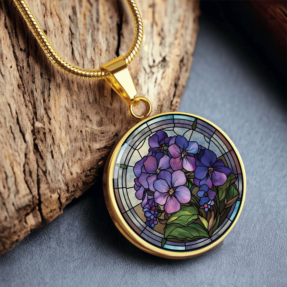 an Elegant gold pendant African Violet Birth Flower Necklace. Violet and purple flowers, green grass, blue, purple, brown background. This jewelry offers personalization with an engraving option for a name.