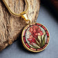 an elegant gold pendant Gladiolus Necklace. The colors are tan/green/blue/purple background, red and pink flowers, green grass, red border. This jewelry offers personalization with an engraving option for a name.
