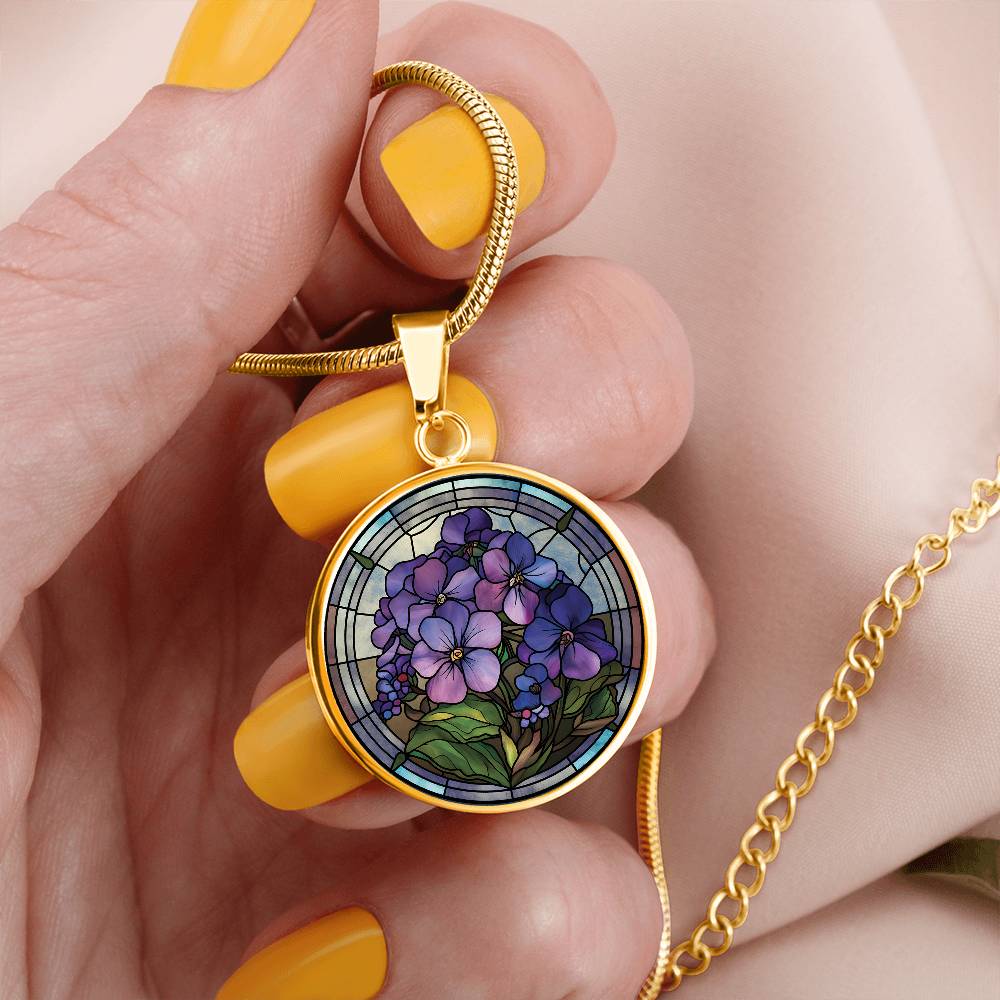 Woman holding an Elegant gold pendant African Violet Birth Flower Necklace. Violet and purple flowers, green grass, blue, purple, brown background. This jewelry offers personalization with an engraving option for a name.