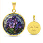 an Elegant gold pendant African Violet Birth Flower Necklace. Violet and purple flowers, green grass, blue, purple, brown background. This jewelry offers personalization with an engraving option for a name.