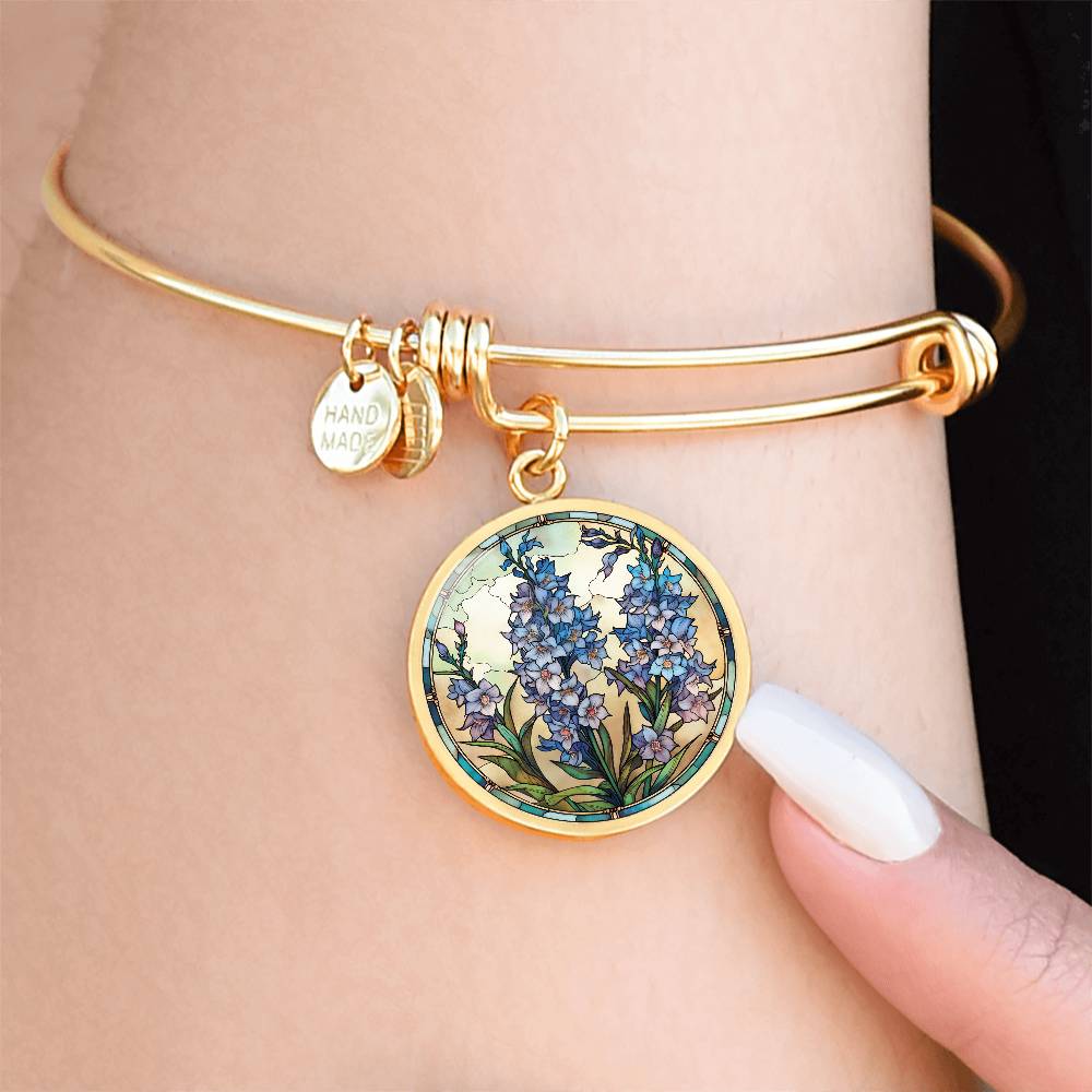 Woman wearing an elegant gold pendant Larkspur Flower Bracelet. The colors are blue, purple, green and tan. This jewelry offers personalization with an engraving option for a name.