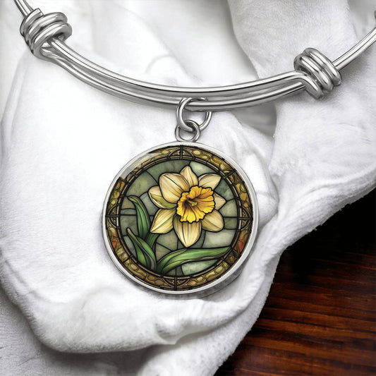 an Elegant silver pendant bracelet featuring a vivid Yellow Daffodil with green grass, set against a tan backdrop with a brown border. This jewelry offers personalization with an engraving option for a name.