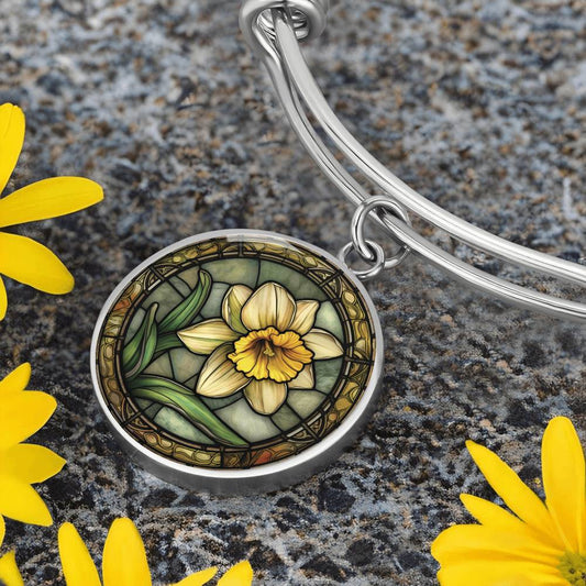 an Elegant silver pendant bracelet featuring a vivid Yellow Daffodil with green grass, set against a tan backdrop with a brown border. This jewelry offers personalization with an engraving option for a name.