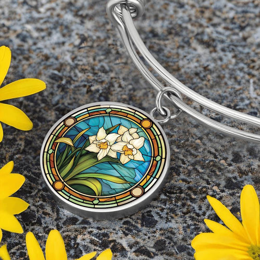 an elegant silver pendant Narcissus Bracelet. Colors are white flowers with yellow centers, green grass, blue background, orange/tan/green border. This jewelry offers personalization with an engraving option for a name.