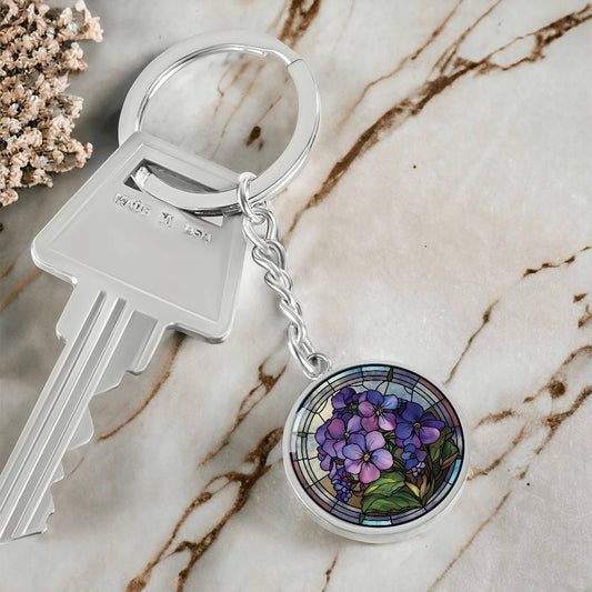 Elegant silver pendant featuring a Purple African Violet Birth Flower Keychain, set against a subtle blue & brown backdrop. This jewelry offers personalization with an engraving option for a name.